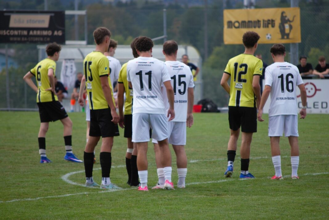 Group of soccer players in yellow and white uniforms standing in a circle on a green field before kickoff. 3 white-jerseyed players with numbers 11 and 15 visible face away from camera.