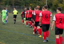 Kampfspiel zwischen Buttisholz und Root Line of soccer players in red jerseys with numbers walking onto a artificial turf field, followed by a neon-yellow goalie and teammates in different kits under a fence.
