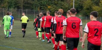 Line of soccer players in red jerseys with numbers walking onto a artificial turf field, followed by a neon-yellow goalie and teammates in different kits under a fence.