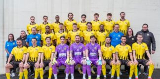 Group portrait of a soccer team in yellow and black kits, with four goalkeepers in purple jerseys seated in front row.