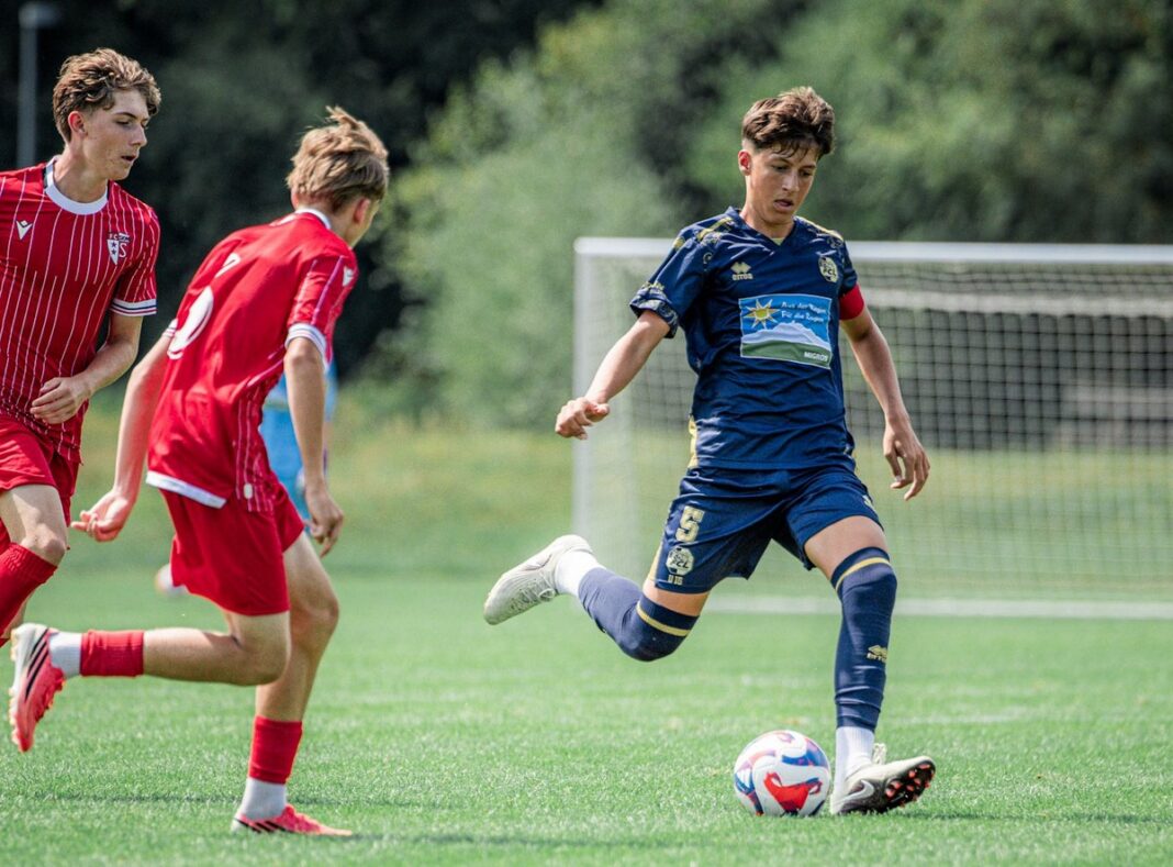 6M5A7166 Youth soccer match: a player in blue controls the ball while two opponents in red chase nearby, goal visible in the background on a sunny field.