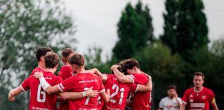 Soccer players in red jerseys huddle together on a green field before a game.