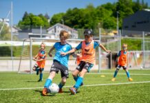MS Sports mit neuem Titelspsonsor für die Football Camps Two young players in blue and orange bibs chase the soccer ball on a sunny field.