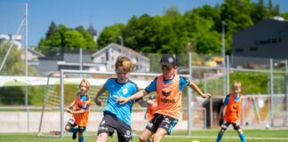 Two young players in blue and orange bibs chase the soccer ball on a sunny field.