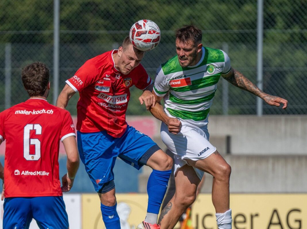 Two soccer players jump to head the ball during a match, one in a red jersey and the other in green-and-white stripes, with a fence backdrop.