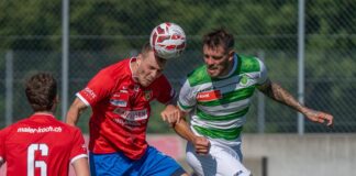 Two soccer players jump to head the ball during a match, one in a red jersey and the other in green-and-white stripes, with a fence backdrop.
