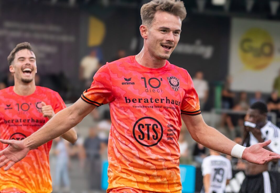 781425501_18627356392011992_4654035224276269661_n Two male soccer players in bright orange jerseys celebrate a goal on the field with a cheering crowd behind them