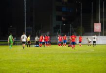 Spielabbruch Emmen gegen Rotkreuz Nighttime soccer match with players in red and white; a group clusters around a fallen teammate on the green field.