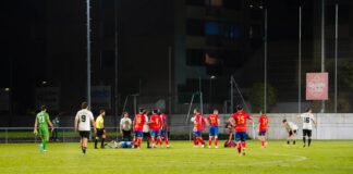 Nighttime soccer match with players in red and white; a group clusters around a fallen teammate on the green field.