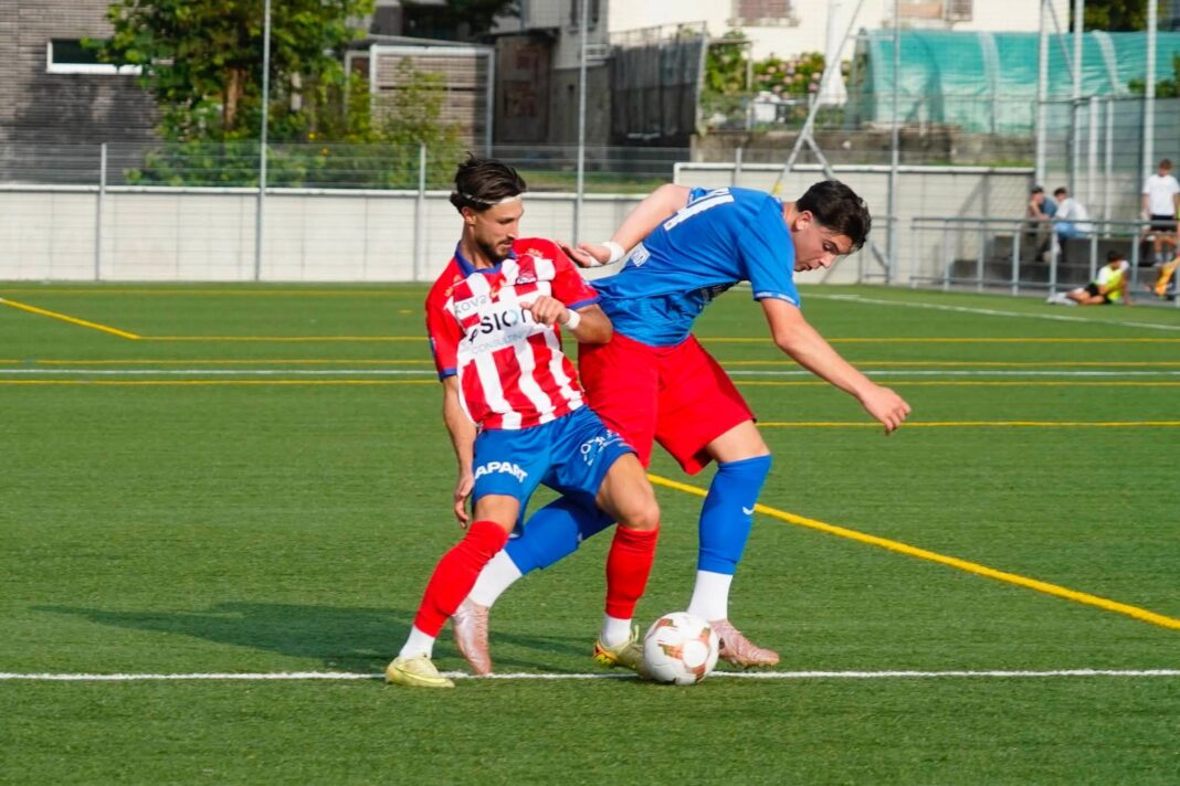 788689477_28490441830552527_474175672539568589_n Two soccer players compete for the ball on a green field; one in a red-and-white striped jersey, the other in blue, near the sideline fence.