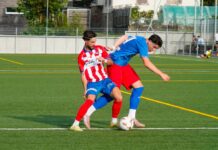 Rotkreuz entscheidet das Nachbarsduell gegen Cham für sich Two soccer players compete for the ball on a green field; one in a red-and-white striped jersey, the other in blue, near the sideline fence.