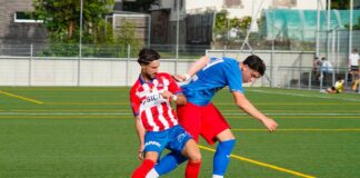 Two soccer players compete for the ball on a green field; one in a red-and-white striped jersey, the other in blue, near the sideline fence.