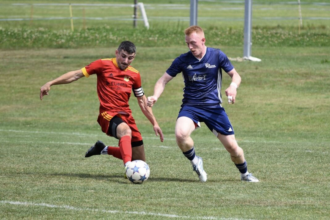 7DEE5552-1D85-40C7-B3E2-58A5BEC21192_1_201_a Two soccer players chase a ball on a grassy field, one in red and the other in blue, competing for possession.
