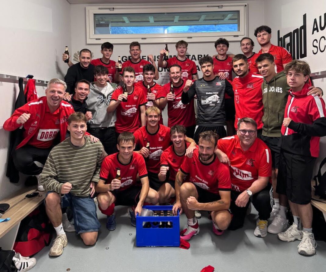 Baar Handball team of about 20 players in red jerseys posing in a locker room, many holding beer bottles and smiling after a game.
