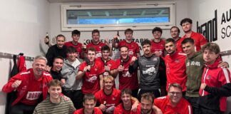 Handball team of about 20 players in red jerseys posing in a locker room, many holding beer bottles and smiling after a game.