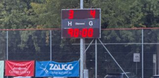 Soccer field with scoreboard showing 3–4 and 90:00; player in orange sits on the turf near banners.