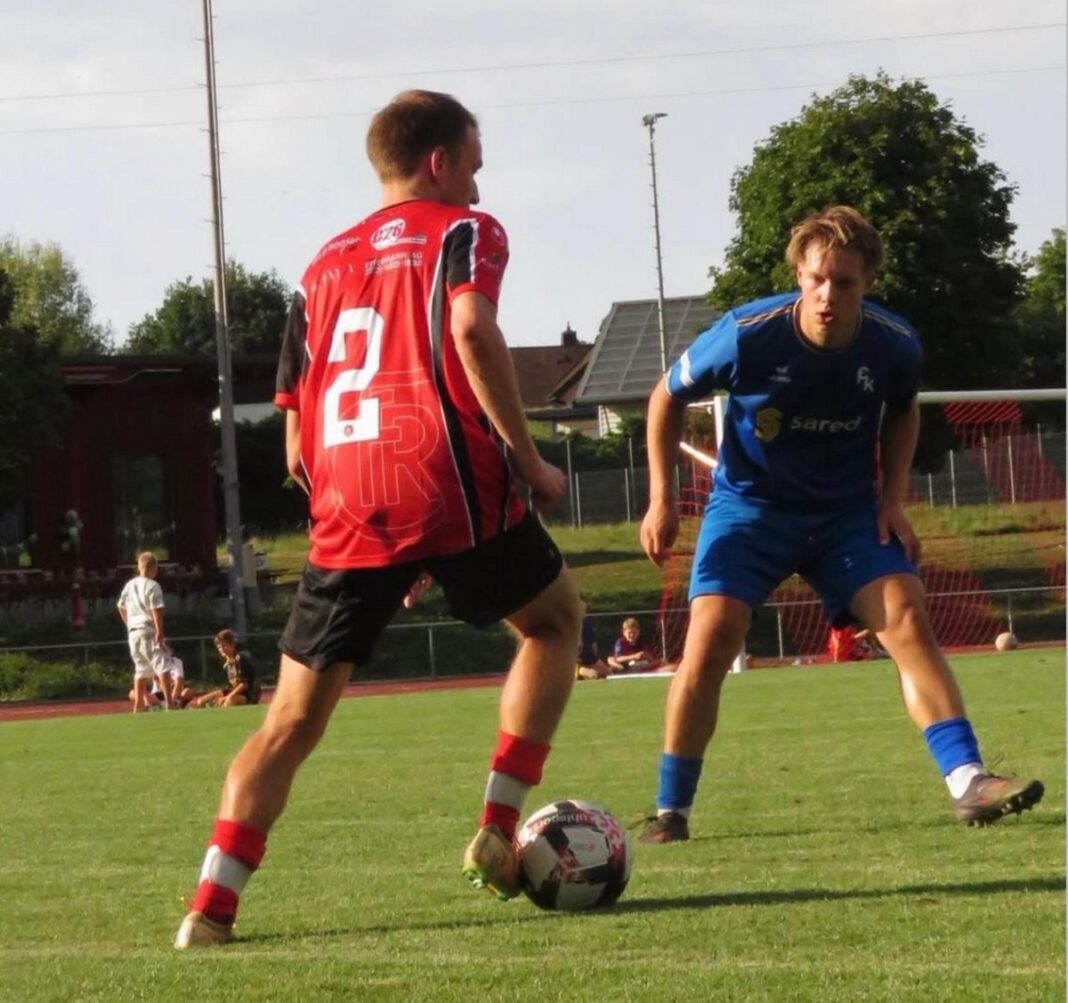 Two soccer players duel for the ball on a grassy field; red-jerseyed attacker (number 2) faces blue-jersey defender near the ball.