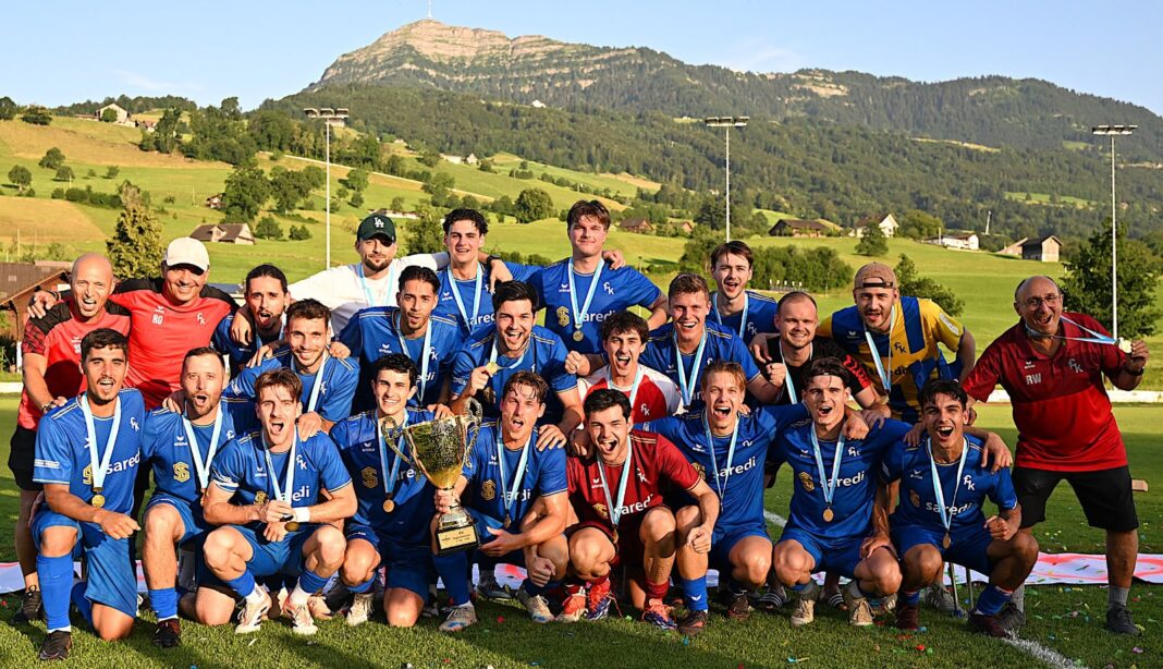 Bild für Bericht Group of men in blue soccer uniforms posing with a large trophy and medals on a sunny grassy field, hills and mountains in the background.