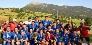 Group of men in blue soccer uniforms posing with a large trophy and medals on a sunny grassy field, hills and mountains in the background.