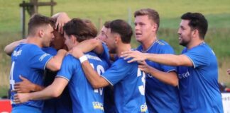 Soccer players in blue jerseys form a huddle, arms around each other in celebration on a field
