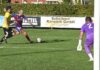 Zwei Standartsituationen entscheiden Duell Brunnen gegen Emmenbrücke Soccer scene: a player in blue and red striped kit kicks the ball while a yellow-jersey opponent approaches on a grassy field, with a purple-clad goalkeeper near the goal in the background