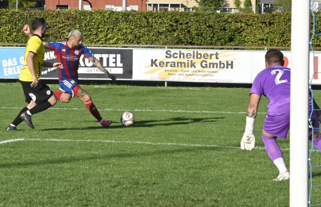 Brunnen-EmmenbrückeDizdarevicGashi Soccer scene: a player in blue and red striped kit kicks the ball while a yellow-jersey opponent approaches on a grassy field, with a purple-clad goalkeeper near the goal in the background