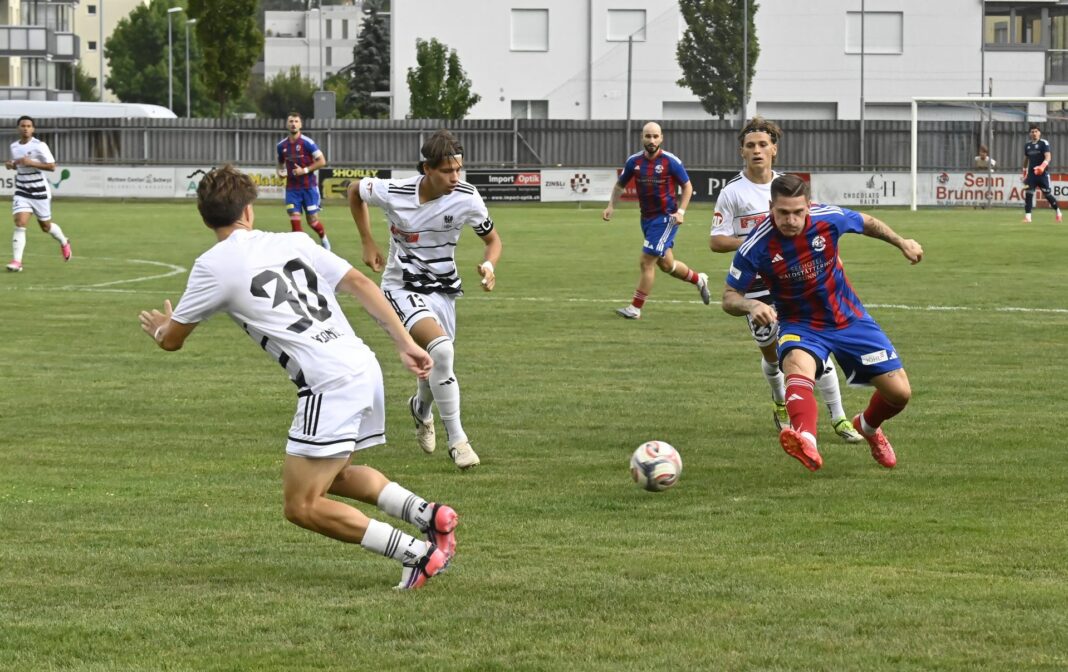 Soccer players in white and blue-red uniforms chase the ball on a green pitch during a match, with a goal and stadium fence in the background bit to the right side area.