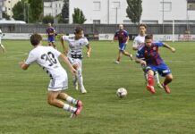 Brunnen schnuppert an der Sensation und wird in der Verlängerung bestraft Soccer players in white and blue-red uniforms chase the ball on a green pitch during a match, with a goal and stadium fence in the background bit to the right side area.