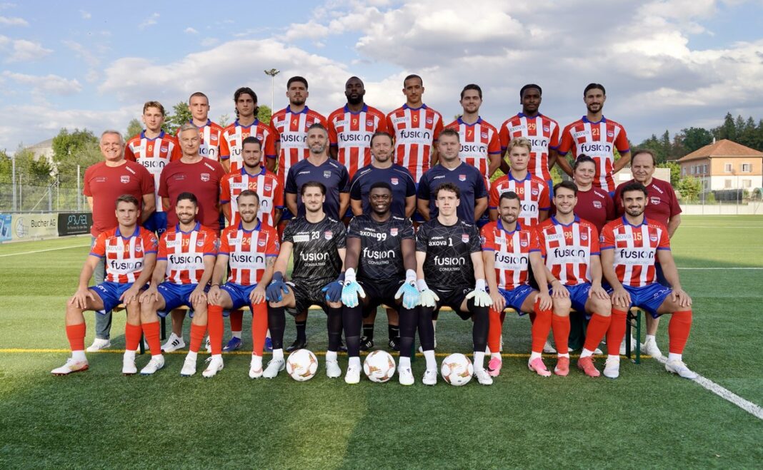 _DSC2813 Group of soccer players and coaches pose for a team photo on a green field, wearing red-and-white striped uniforms and blue shorts on a sunny day.