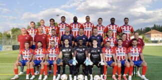 Group of soccer players and coaches pose for a team photo on a green field, wearing red-and-white striped uniforms and blue shorts on a sunny day.