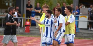 Soccer players in white and blue striped kits celebrate near the sideline, gesturing and smiling at a coach