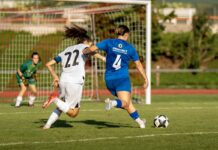 Küssnachter Frauen starten mit Sieg in die neue Nationalliga-B-Saison Two female soccer players chase the ball toward the goal; blue jersey #4 and white jersey #22 compete, with a green-clad goalkeeper in the background.
