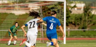 Two female soccer players chase the ball toward the goal; blue jersey #4 and white jersey #22 compete, with a green-clad goalkeeper in the background.