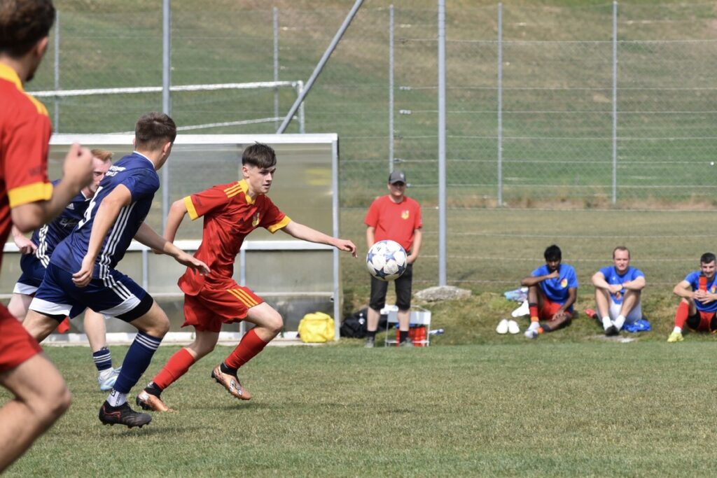 F21C7932-D6EF-41A1-8C89-46CE909B5239_1_201_a-1024x683 Youth soccer match: a player in red chases a ball near the goal area with blue-clad opponents close behind on a grassy field.
