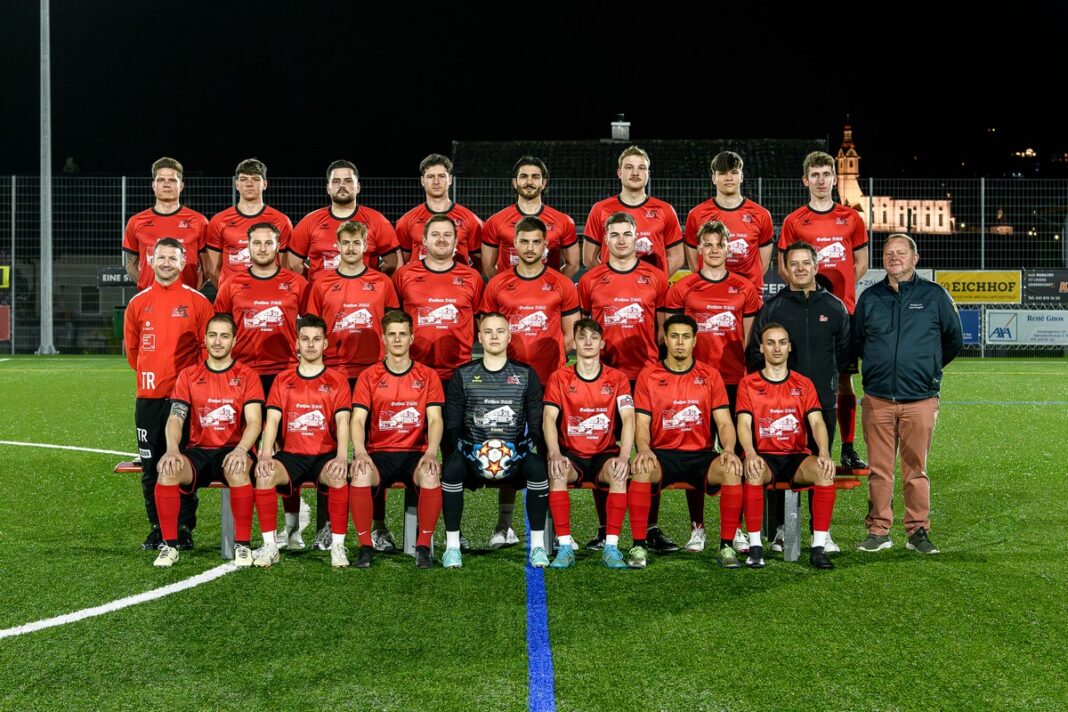 FCS_2_Web-1-1 Group photo of a red-jersey soccer team posing on a night field, players arranged in rows with a goalkeeper holding a ball in the center.