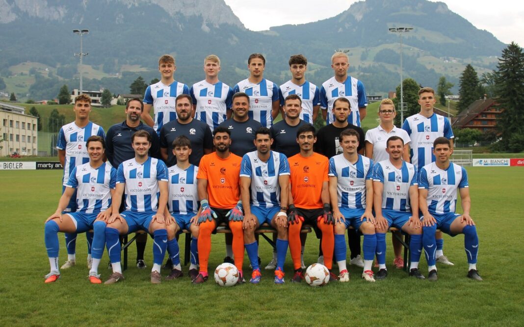 Foto FC Ibach in der Vorbereitung (1) Group photo of a soccer team on a grass field with mountains in the background, players in blue-and-white striped jerseys and orange goalkeepers in front row.