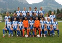 Saisonvorschau: Der FC Ibach startet mit viel Elan in die neue Saison Group photo of a soccer team on a grass field with mountains in the background, players in blue-and-white striped jerseys and orange goalkeepers in front row.