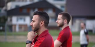Two men in red soccer jerseys stand on a field; foreground man with hand on chin and tattooed arm.