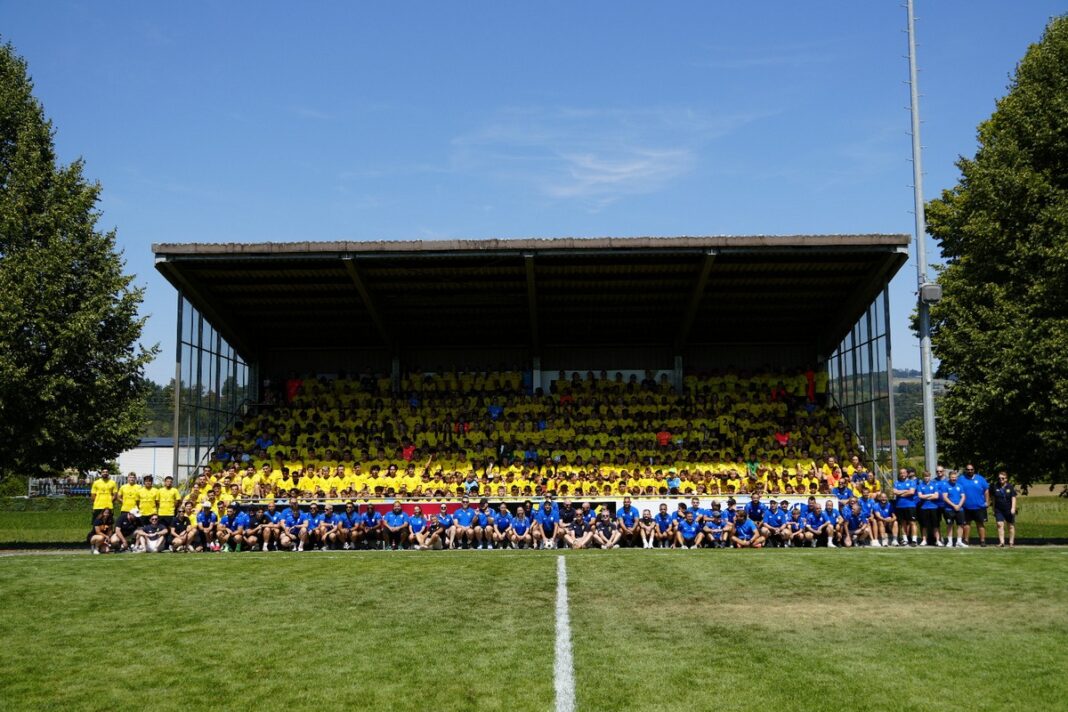 Large group photo in a stadium: many people in yellow shirts seated in the bleachers, with a blue-shirt group on the grass in front, under a clear blue sky.