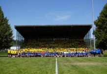 Gelungener Kick-Off in die neue Saison beim FC Baar Large group photo in a stadium: many people in yellow shirts seated in the bleachers, with a blue-shirt group on the grass in front, under a clear blue sky.