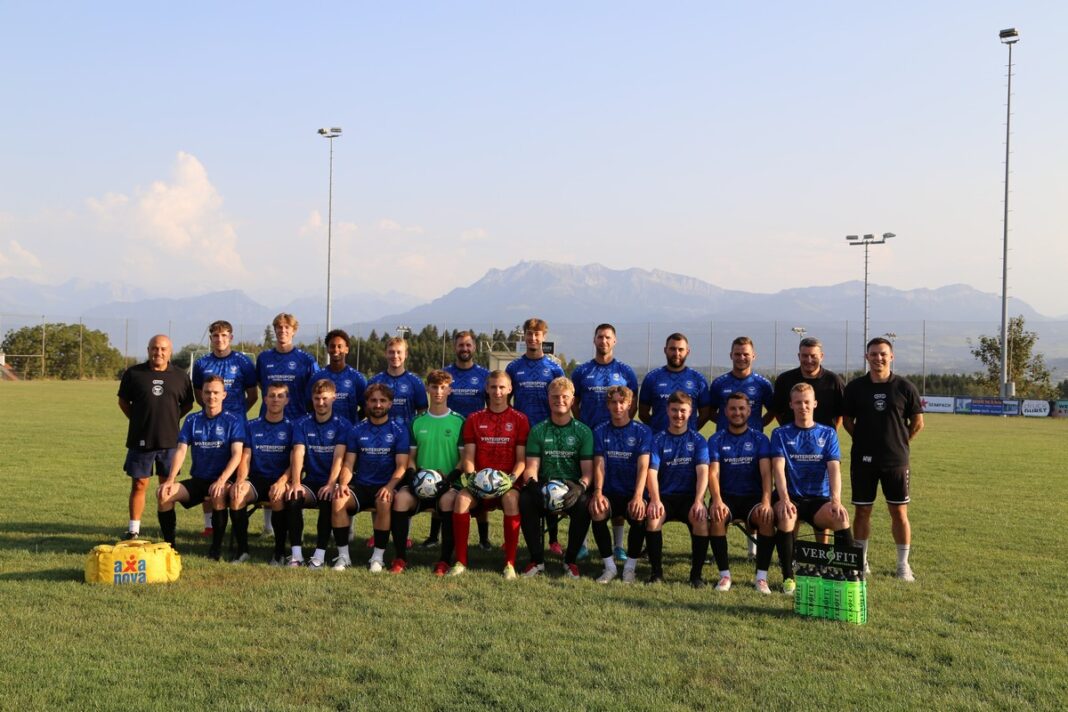 HSVI Group of soccer players on a grassy field posing for a team photo; 22 players in blue uniforms with two goalkeepers in red and green, mountain backdrop behind.