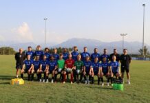 Starker Saisonstart: Hildisrieden lässt Adligenswil keine Chance Group of soccer players on a grassy field posing for a team photo; 22 players in blue uniforms with two goalkeepers in red and green, mountain backdrop behind.