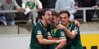 Three soccer teammates in green jerseys celebrate a goal together on the field, arms around each other, smiles on their faces in front of a stadium crowd.