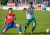 Cup-Derby im Eizmoos: SC Cham empfängt den SC Kriens Two soccer players compete for the ball on a sunny grass field, one in red jersey and blue shorts sliding while the other in white shirt and green shorts reaches for the ball behind him.