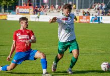 Cup-Derby im Eizmoos: SC Cham empfängt den SC Kriens Two soccer players compete for the ball on a sunny grass field, one in red jersey and blue shorts sliding while the other in white shirt and green shorts reaches for the ball behind him.