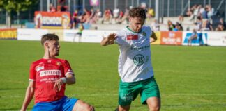 Two soccer players compete for the ball on a sunny grass field, one in red jersey and blue shorts sliding while the other in white shirt and green shorts reaches for the ball behind him.