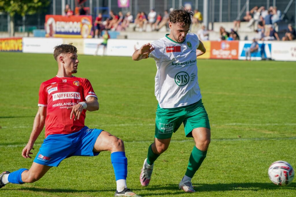 MRG01797-1024x683-1 Two soccer players compete for the ball on a sunny grass field, one in red jersey and blue shorts sliding while the other in white shirt and green shorts reaches for the ball behind him.