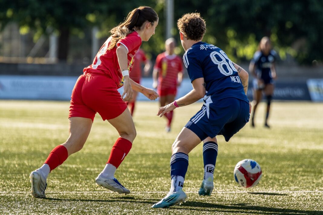 Nicht alle Duelle konnten gewonnen werden Two female soccer players in red and blue uniforms chase a ball on a sunny turf field.