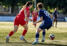 Küssnacht verspielt Führung und unterliegt Carouge mit 2:3 Two female soccer players in red and blue uniforms chase a ball on a sunny turf field.