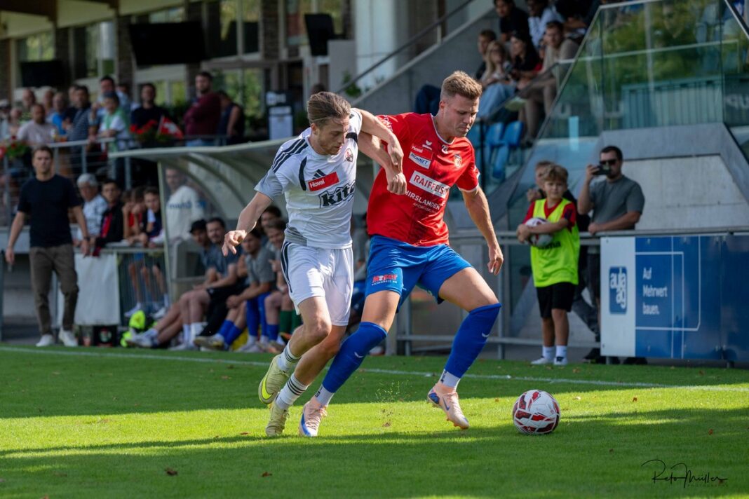 SC Cham_GrandSaconnex-4 Two soccer players battle for the ball on a sunny field, white kit against red kit, with spectators in the stands behind them.